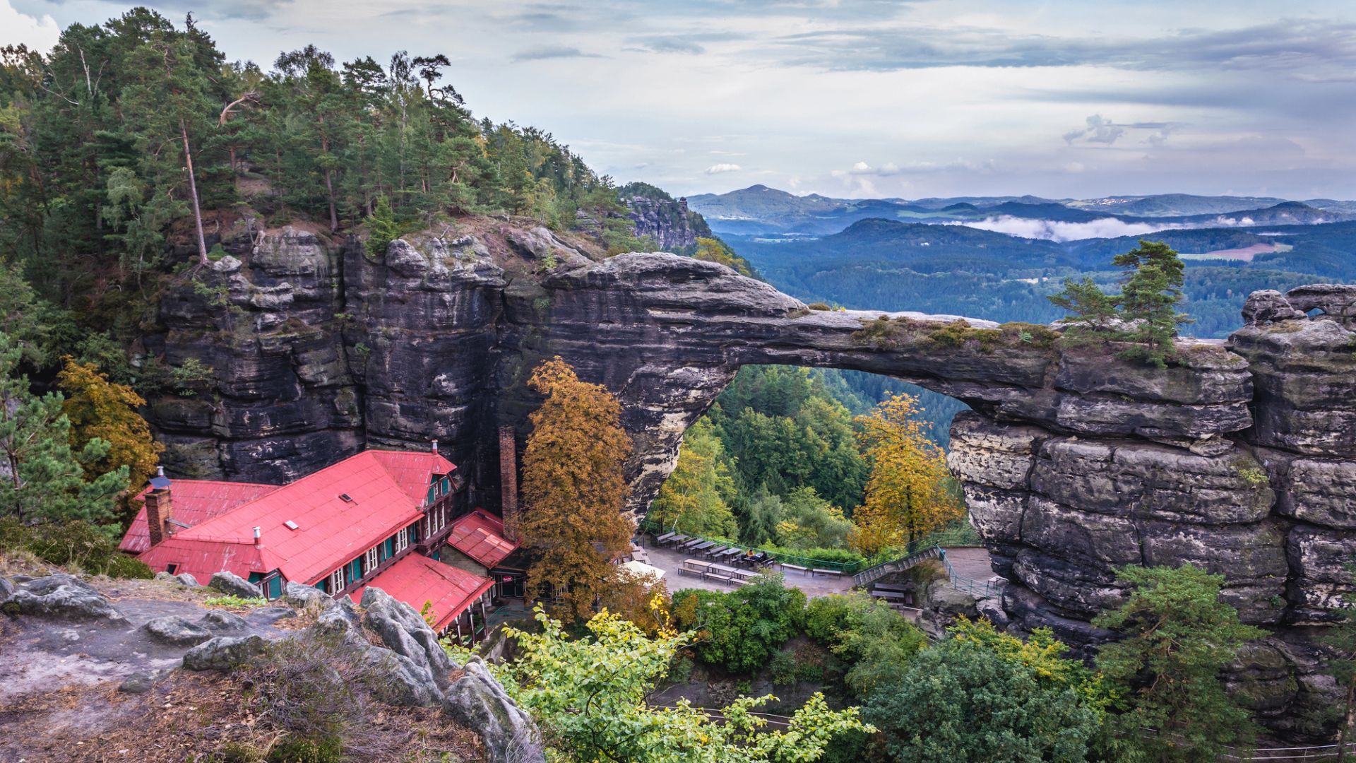 Rock arch bridge with a red-roofed building below, surrounded by trees and rolling hills in the background.