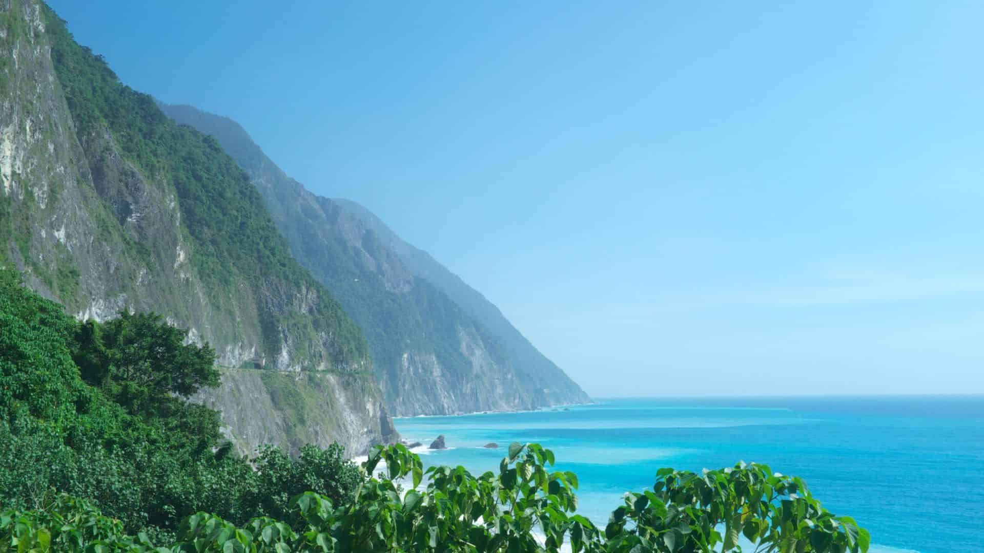 Turquoise ocean waves meet lush green cliffs under a clear blue sky on a sunny day.