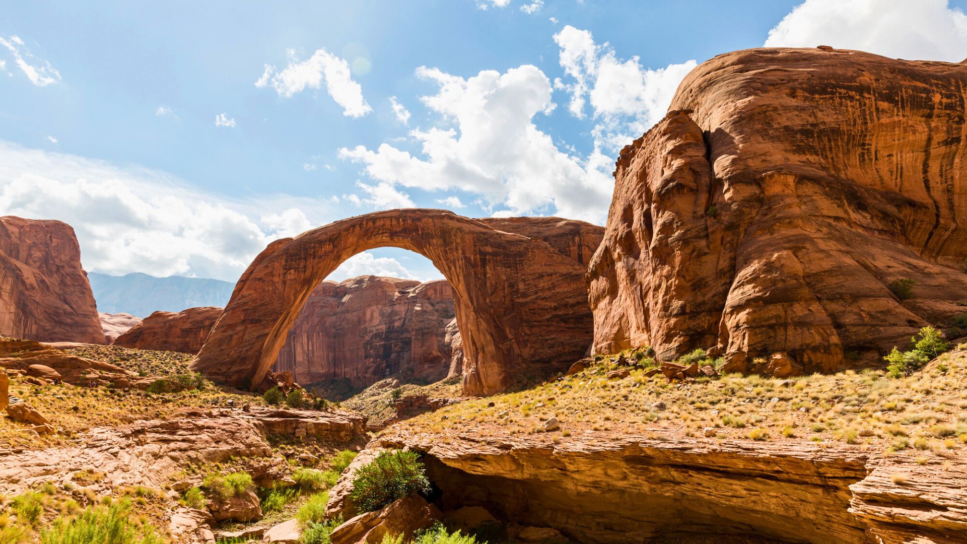 A natural sandstone arch stretches between red rock cliffs under a partly cloudy blue sky in a desert landscape.