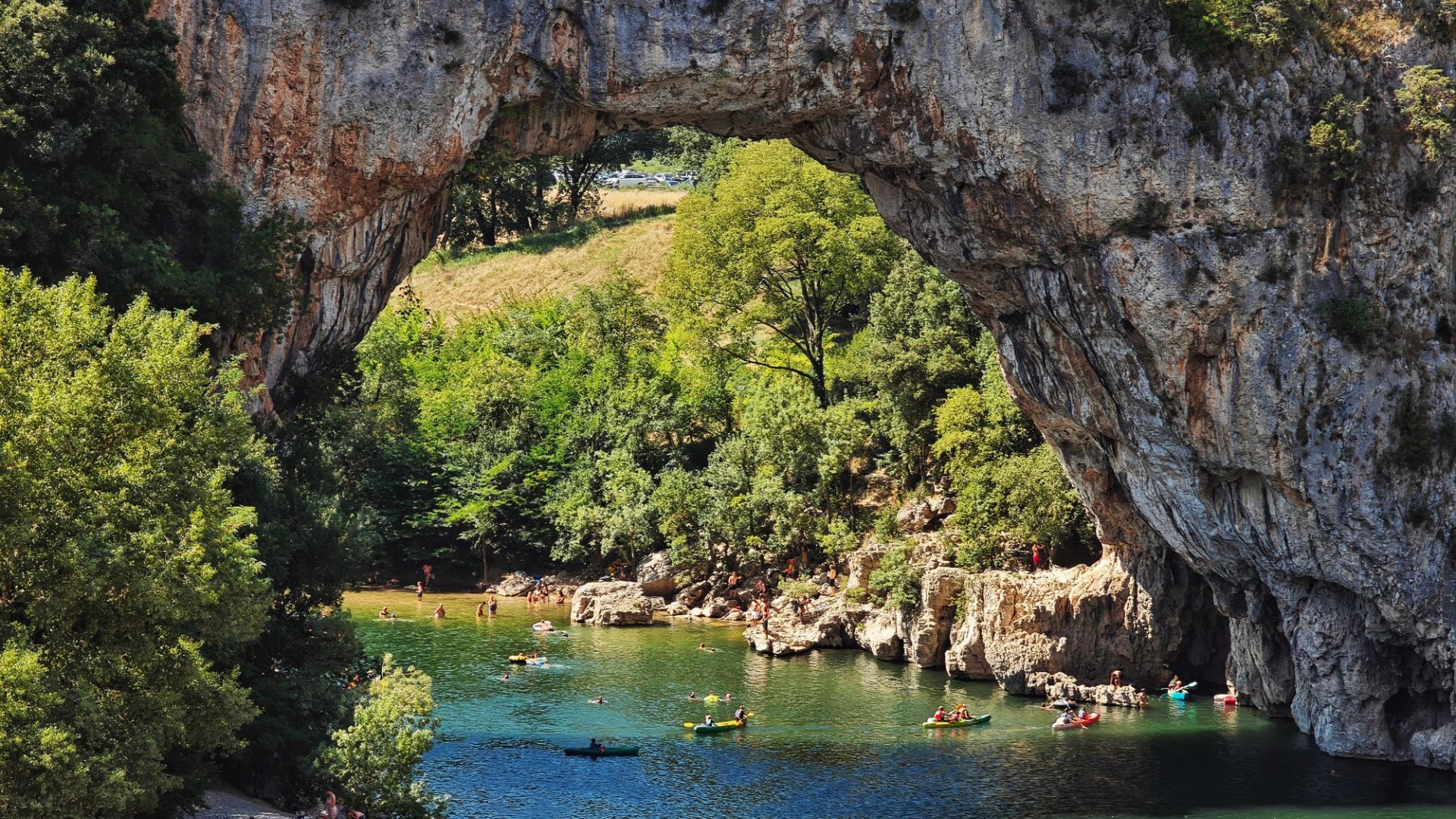 People kayak and swim beneath a large natural stone arch over a river surrounded by greenery.