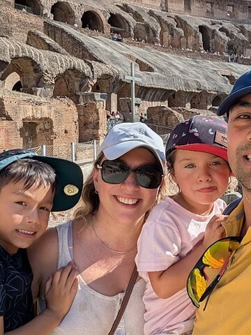 A family of four smiles for a selfie in front of the Colosseum in Rome on a sunny day, capturing the joy of exploring Rome with kids.