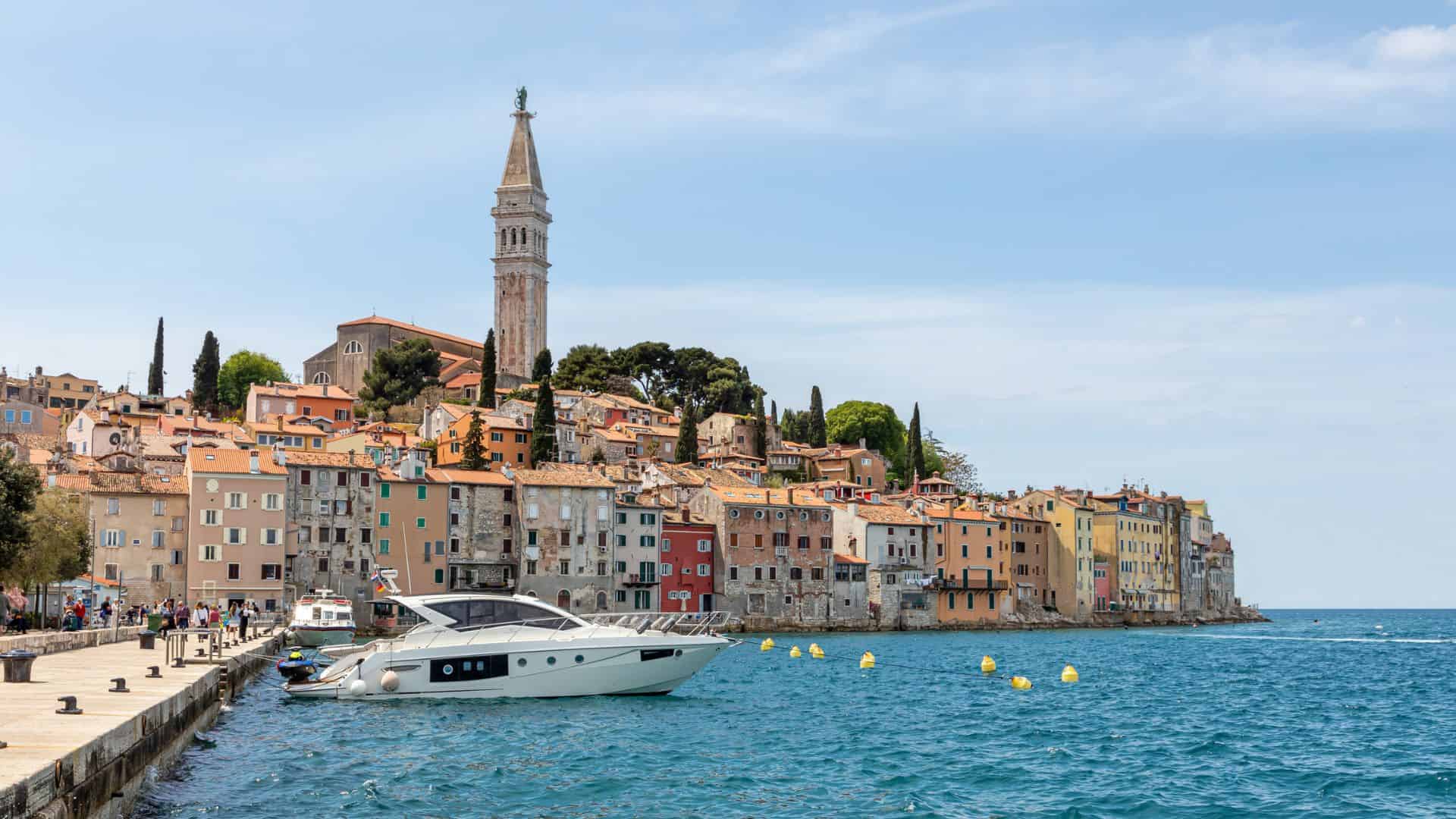Colorful seaside buildings and a tall church tower overlook a marina with boats on a sunny day.