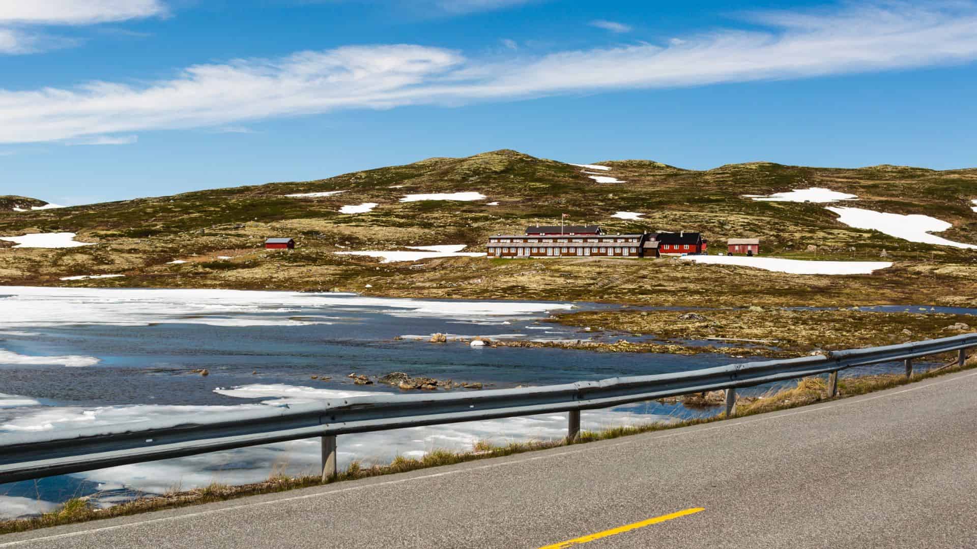 Mountain inn near a partially frozen lake, with snow patches and a winding road in the foreground under a blue sky.