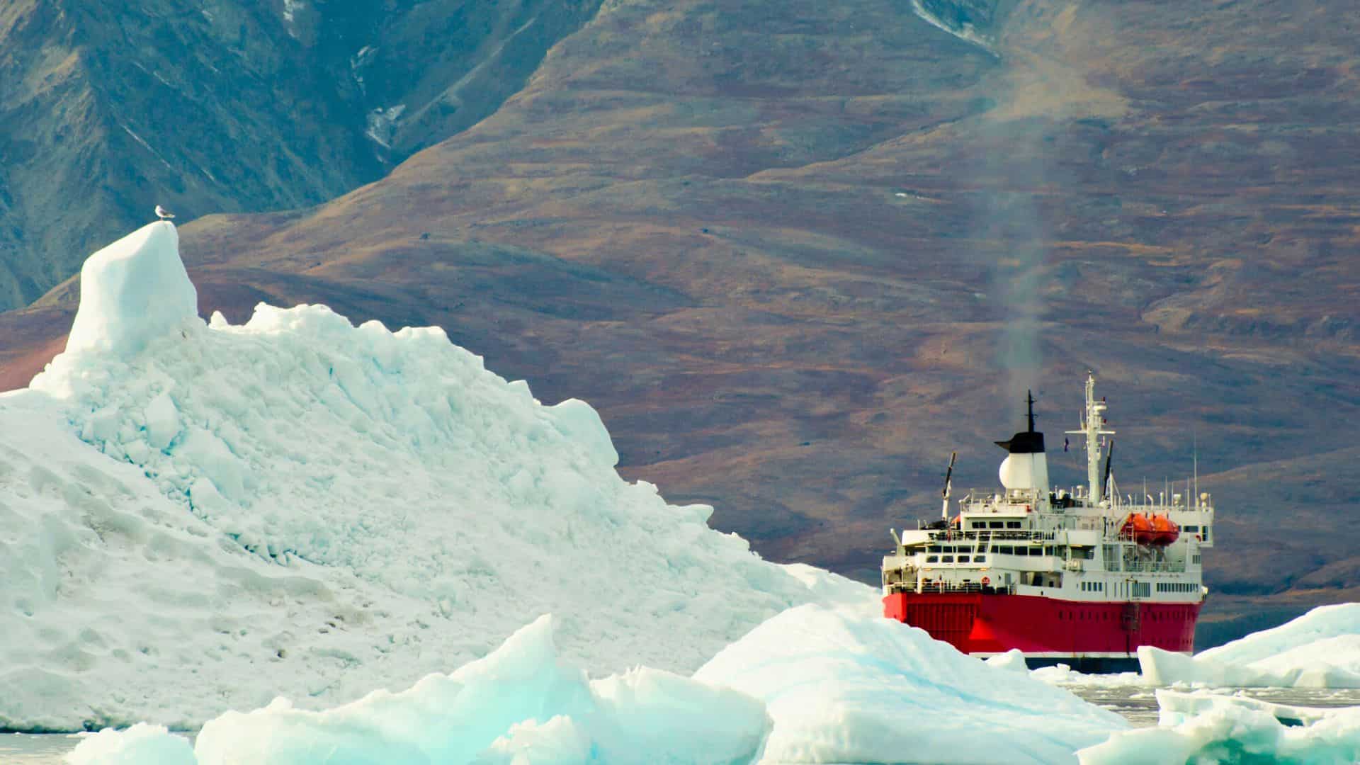 A red and white ship navigates through icy waters near large icebergs, with mountains in the background.