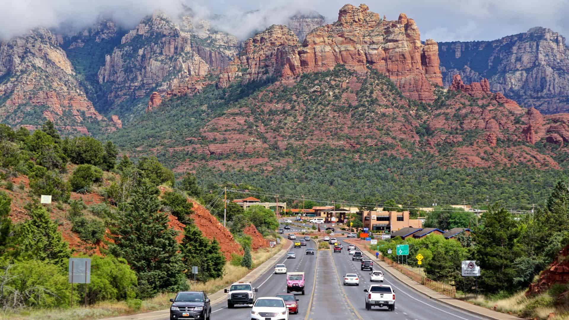 Cars driving on a road toward red rock mountains and green trees under a partly cloudy sky.