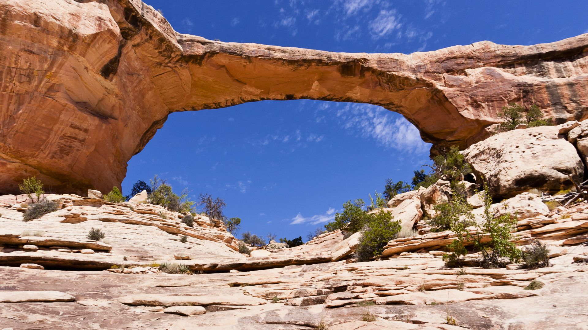 A natural stone arch rises over rocky terrain under a bright blue sky with scattered clouds.