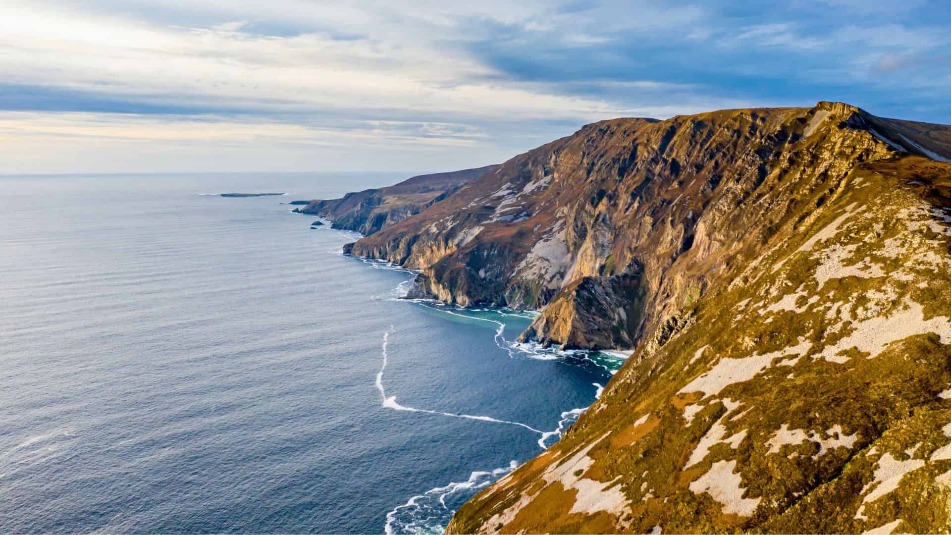 Aerial view of steep sea cliffs meeting the ocean under a cloudy blue sky.