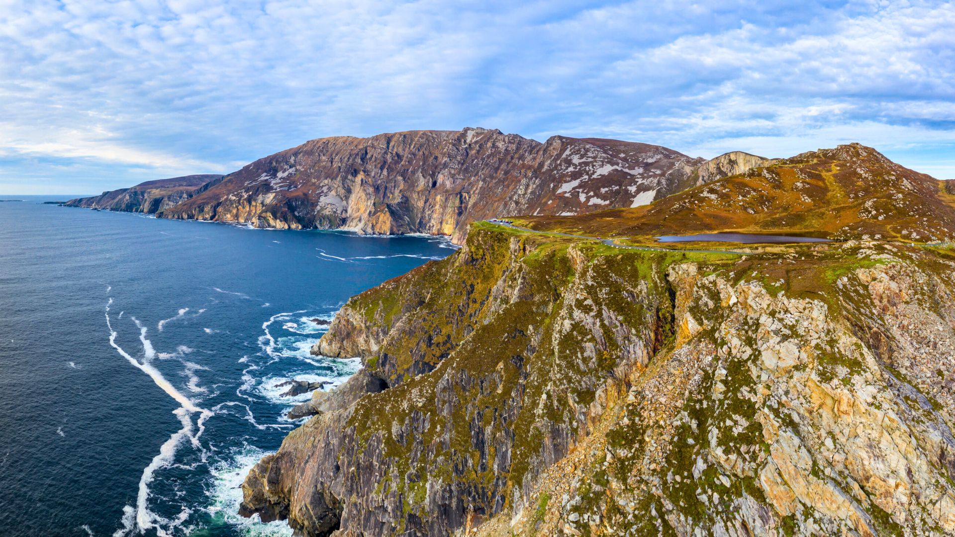 Cliffs with rugged rocks beside deep blue ocean under a partly cloudy sky, with grass on the cliff tops.