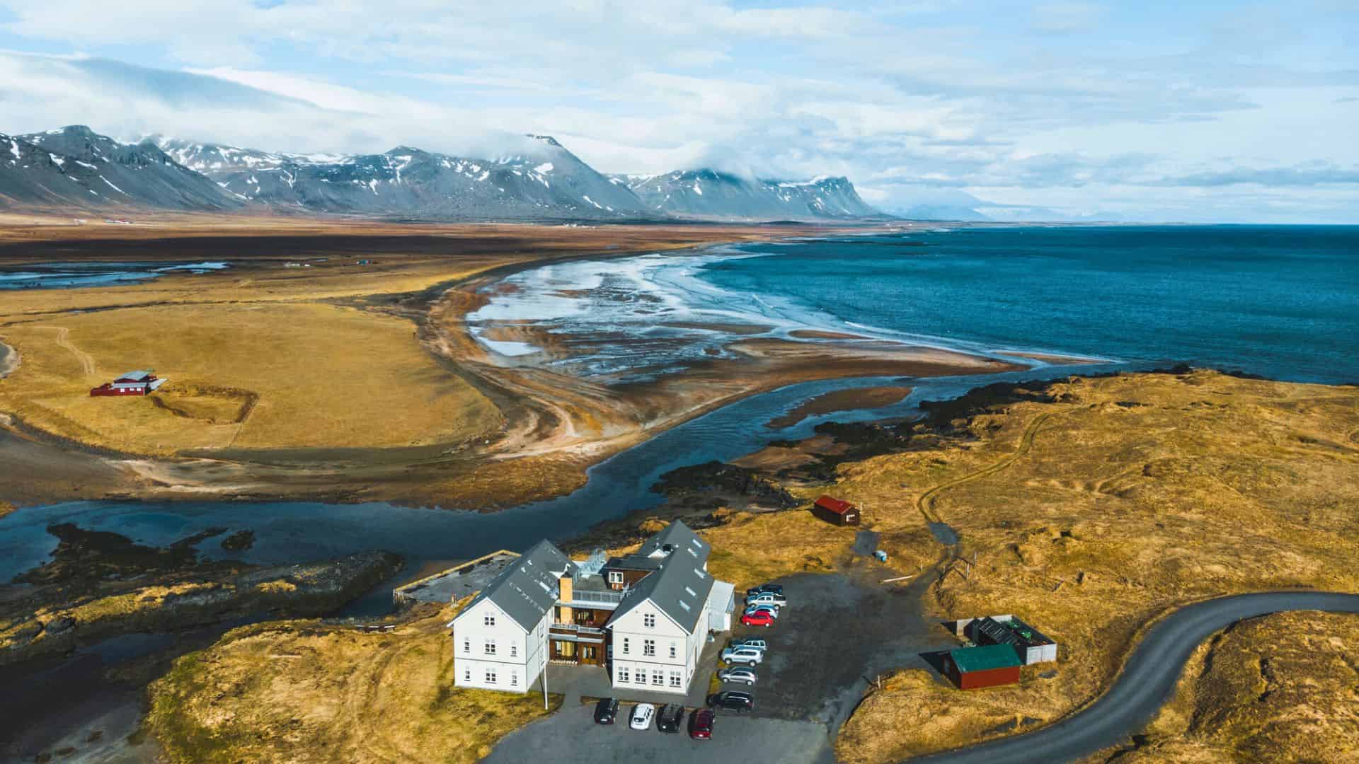 Aerial view of a coastal building with cars, mountains, and shoreline under a partly cloudy sky.