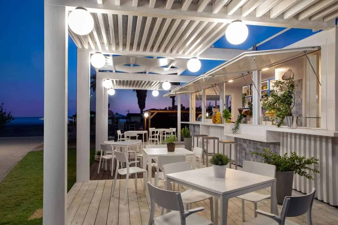 Outdoor cafe with white tables and chairs under pergola lighting, near the beach at sunset, just moments from Parco della Gallinara.