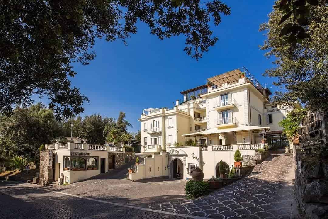 Elegant cream-colored hotel with balconies and a restaurant, surrounded by greenery under a clear blue sky—an ideal retreat for 4 days in Castel Gandolfo.
