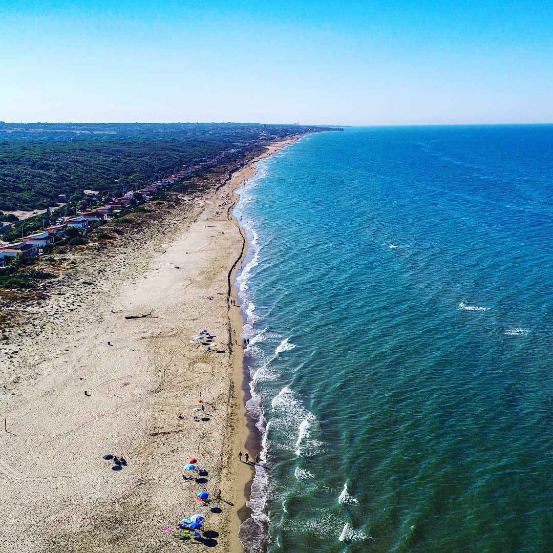 Aerial view of a sandy beach at Parco della Gallinara with umbrellas, people, and calm blue ocean waves under a clear sky.