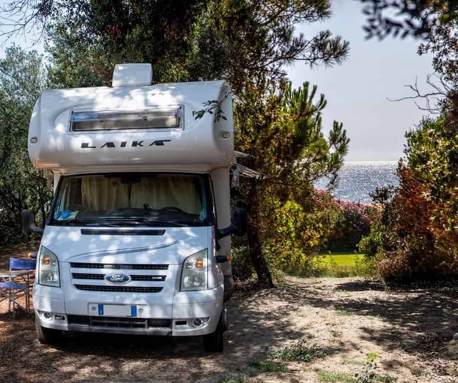 A white campervan is parked among trees near the coast at Parco della Gallinara, with the sea visible in the background.