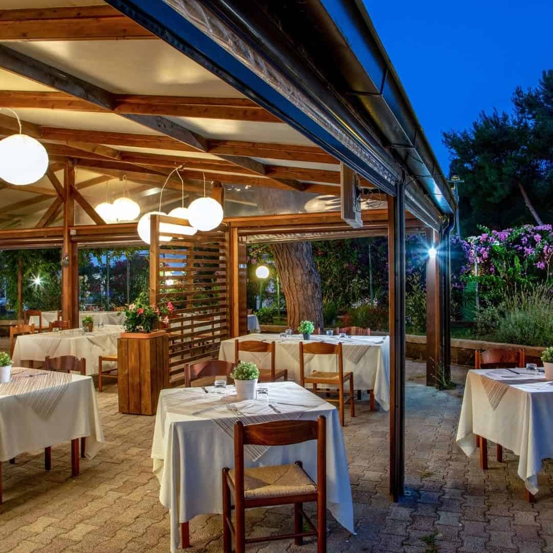 Outdoor restaurant with wooden tables, white tablecloths, and hanging lights in a garden setting at dusk, inspired by the serene atmosphere of Parco della Gallinara.