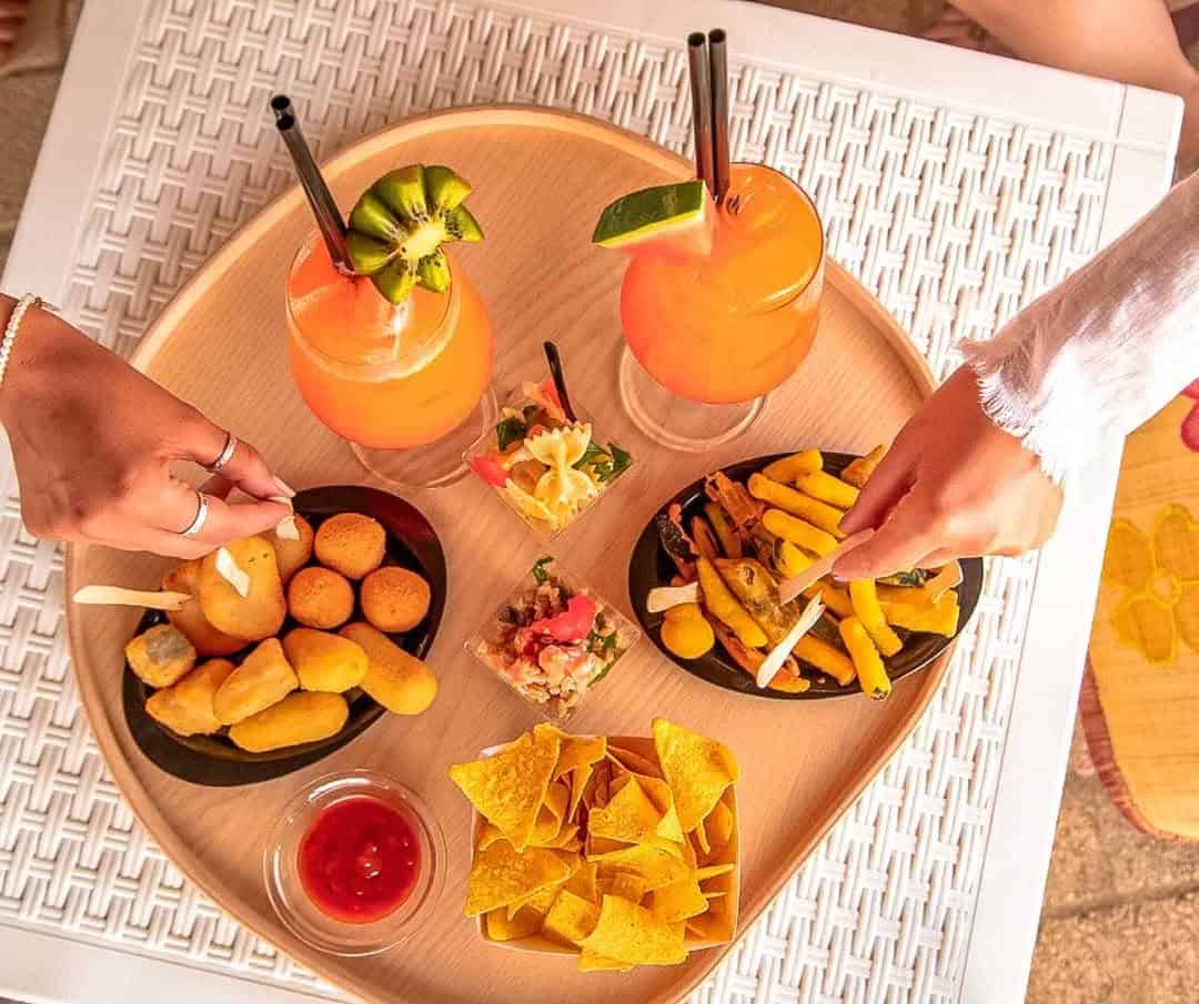 Two hands take snacks from a tray with cocktails, chips, fries, and assorted finger foods on a table at Parco della Gallinara.