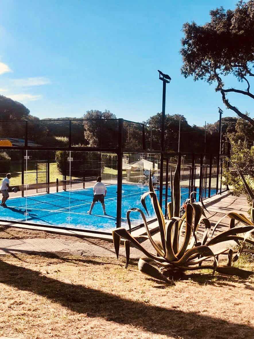 Two people play padel tennis on a blue court outdoors at Parco della Gallinara, surrounded by trees and plants on a sunny day.