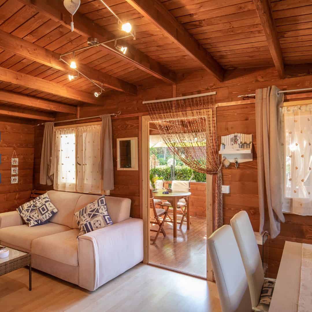 Cozy wooden cabin living room with beige sofa, decorative pillows, and a view of the patio with outdoor seating&mdash;perfect for relaxing after a day exploring Parco della Gallinara.