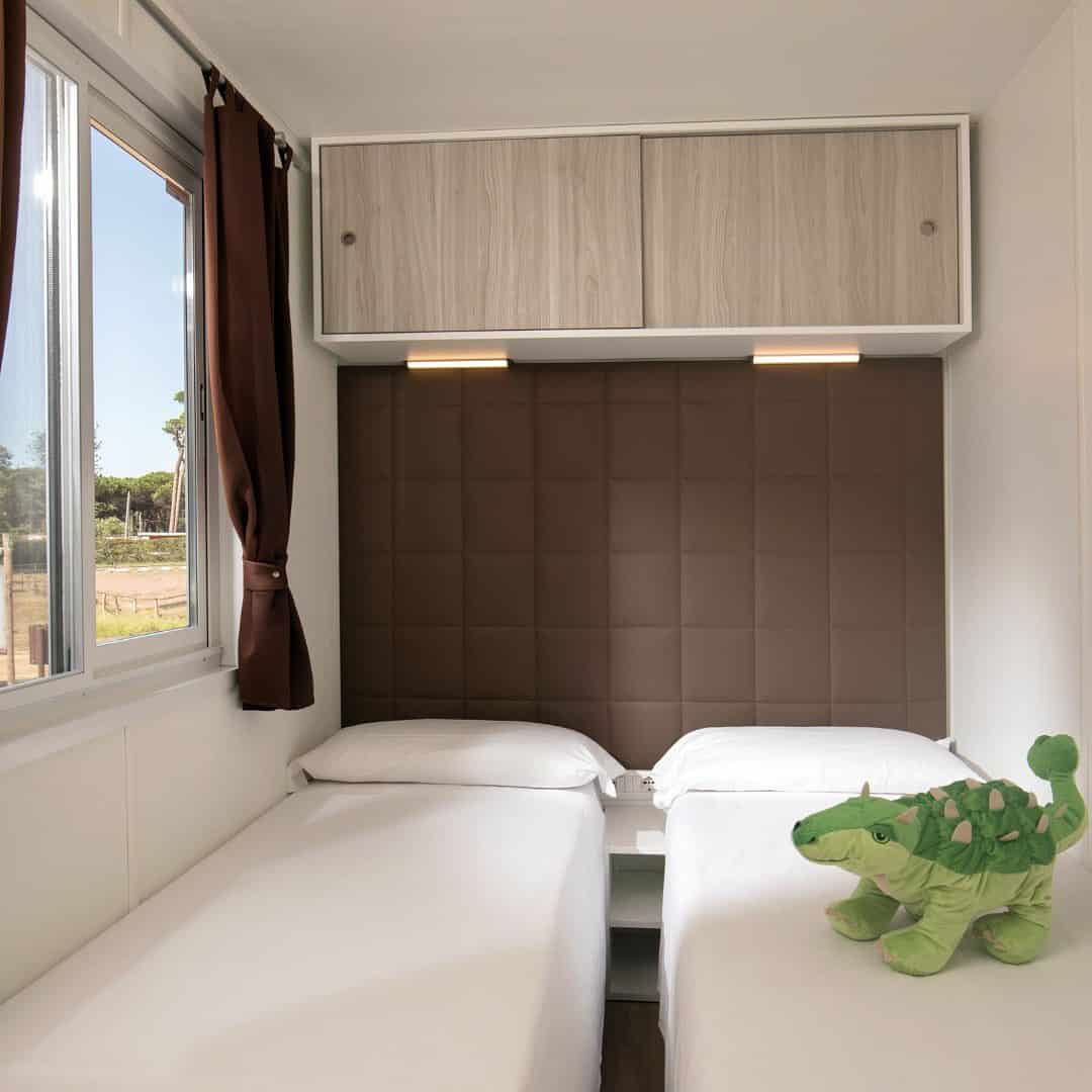 Two single beds with white sheets, a green stuffed toy turtle, and a window with brown curtains create a cozy space, perfect for relaxing after exploring Parco della Gallinara.