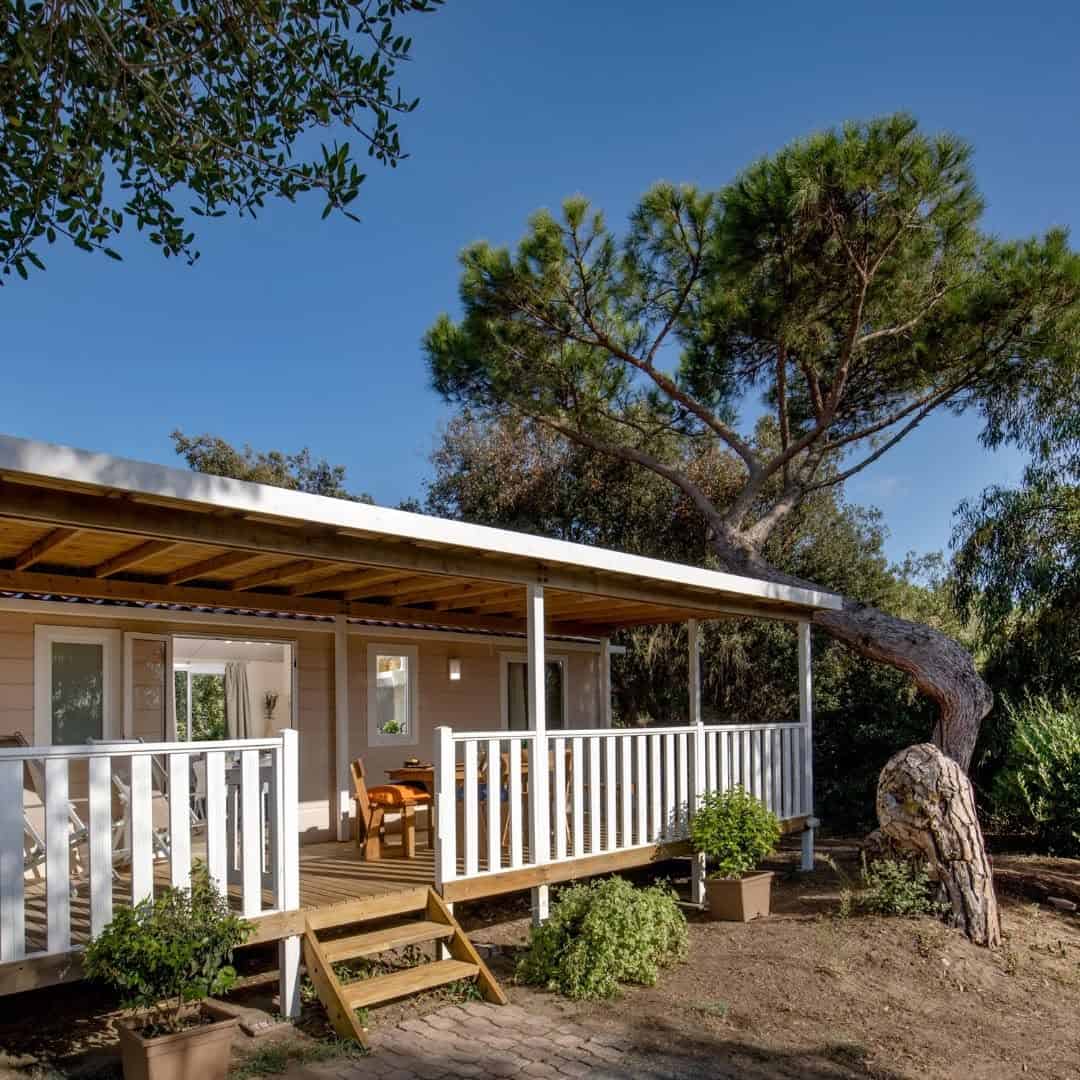 A wooden cabin with a porch and white railing, nestled in the lush greenery of Parco della Gallinara, surrounded by trees under a clear blue sky.