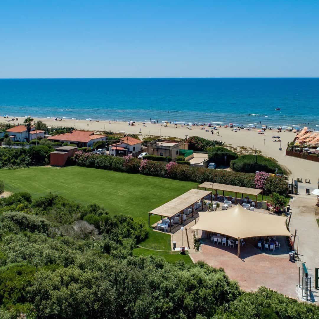 Aerial view of Parco della Gallinara beach resort with green lawns, shaded seating, and people by the sea on a sunny day.