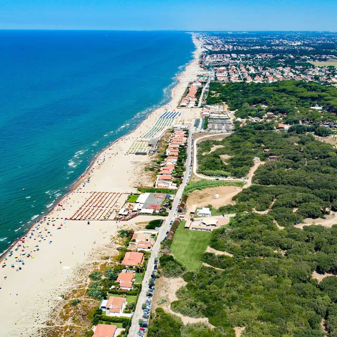 Aerial view of a coastline with sandy beach, blue sea, houses, and green trees along the shore near Parco della Gallinara.