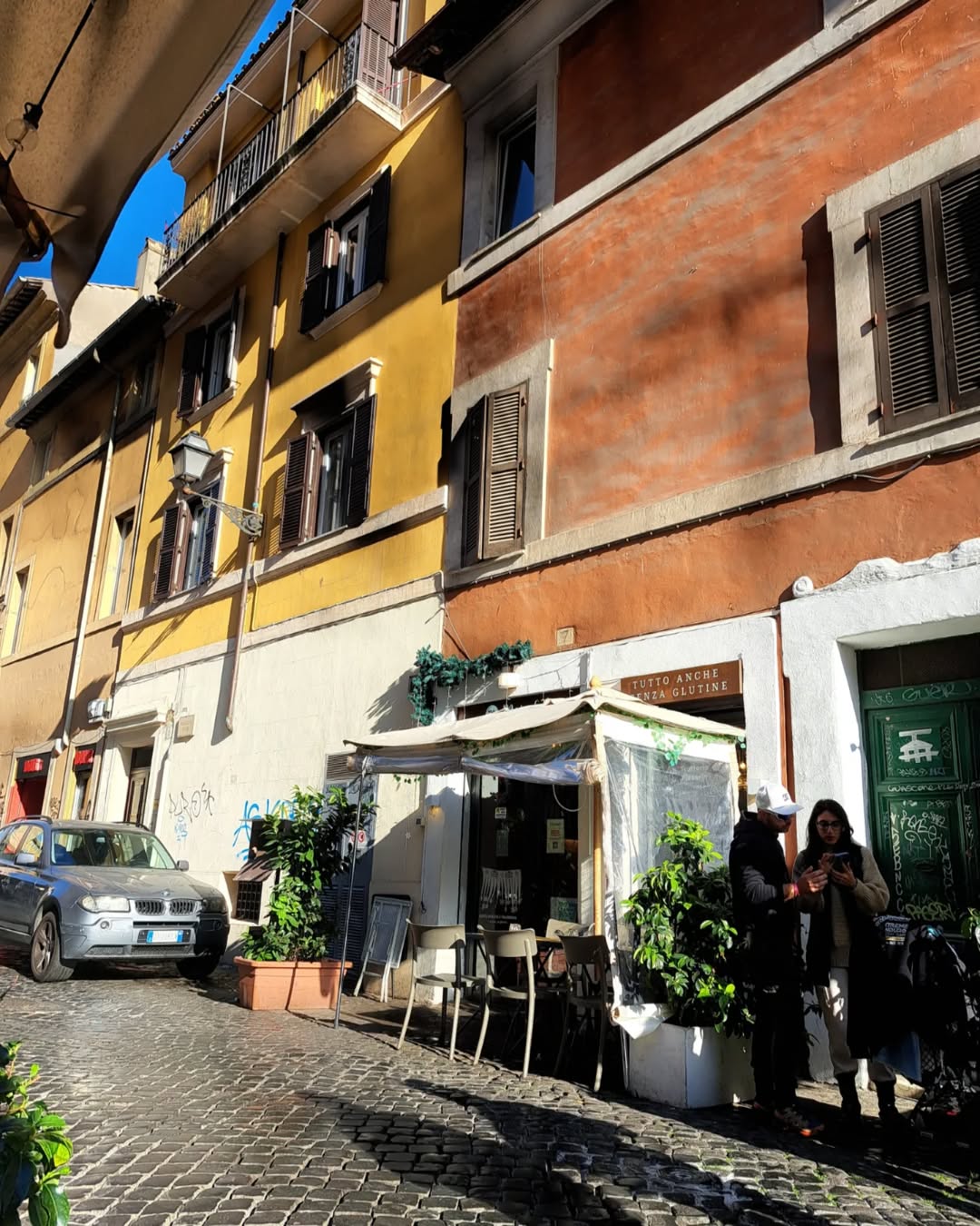 Sunny street with colorful buildings, a parked car, café seating, and two people chatting outside—capturing the charm of discovering where to eat in Rome.