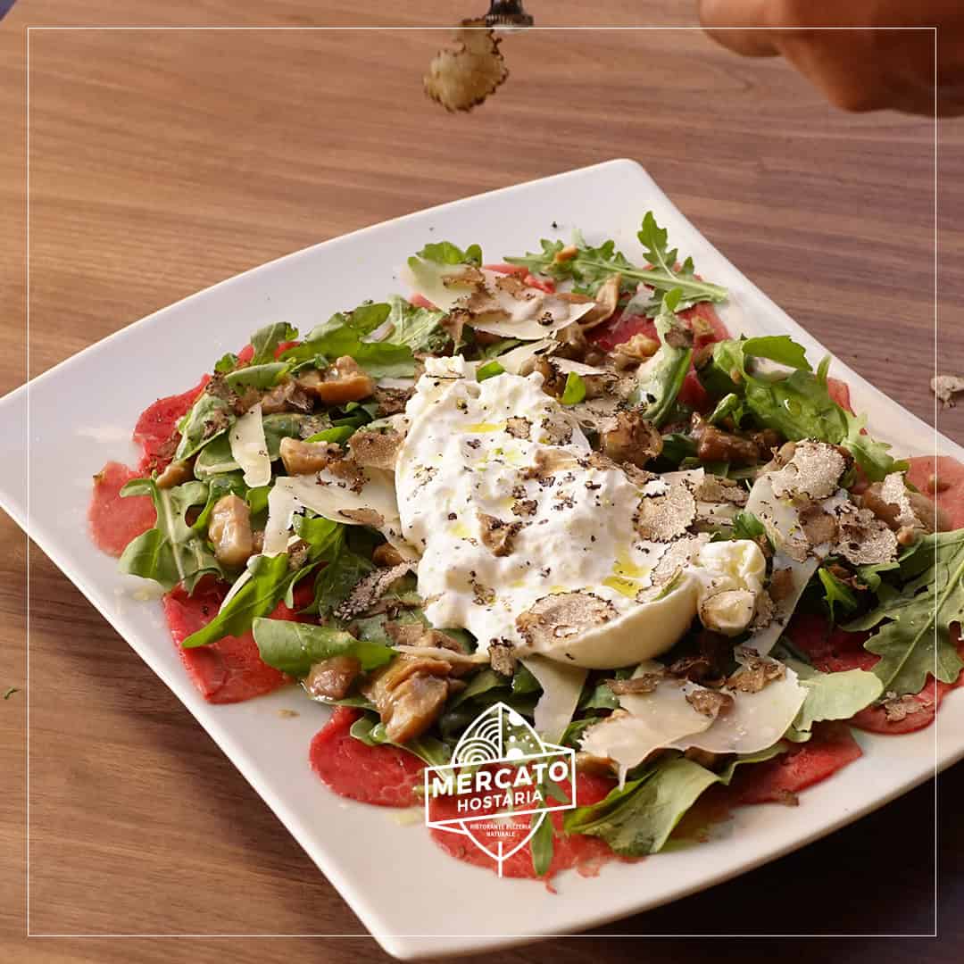 A plate of beef carpaccio with burrata, arugula, walnuts, shaved truffle, and cheese on a wooden table—an ideal dish for those seeking where to eat in Rome.