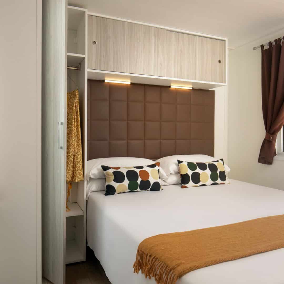 Modern bedroom with a double bed, brown headboard, colorful pillows, and an open closet with hanging clothes&mdash;a relaxing retreat inspired by the natural beauty of Parco della Gallinara.