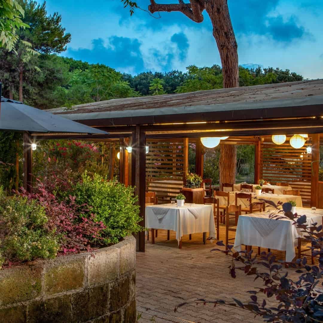 Outdoor restaurant patio at dusk with wooden tables, chairs, and hanging lights, surrounded by lush greenery reminiscent of Parco della Gallinara.