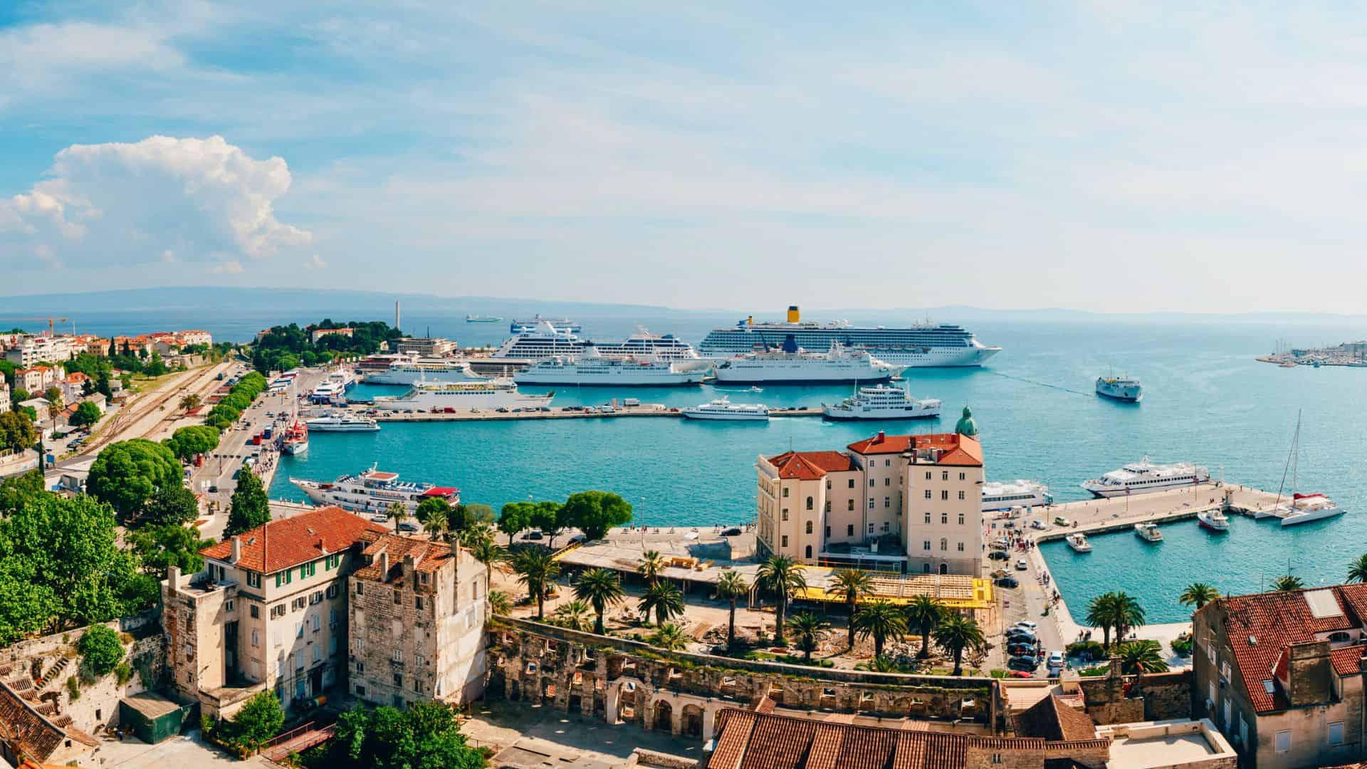 Aerial view of a coastal city harbor with cruise ships, boats, and historic buildings by the sea.
