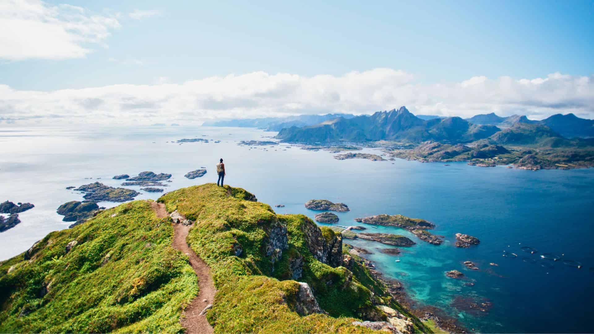 A person stands on a grassy hilltop overlooking blue ocean and distant mountains under a clear sky.