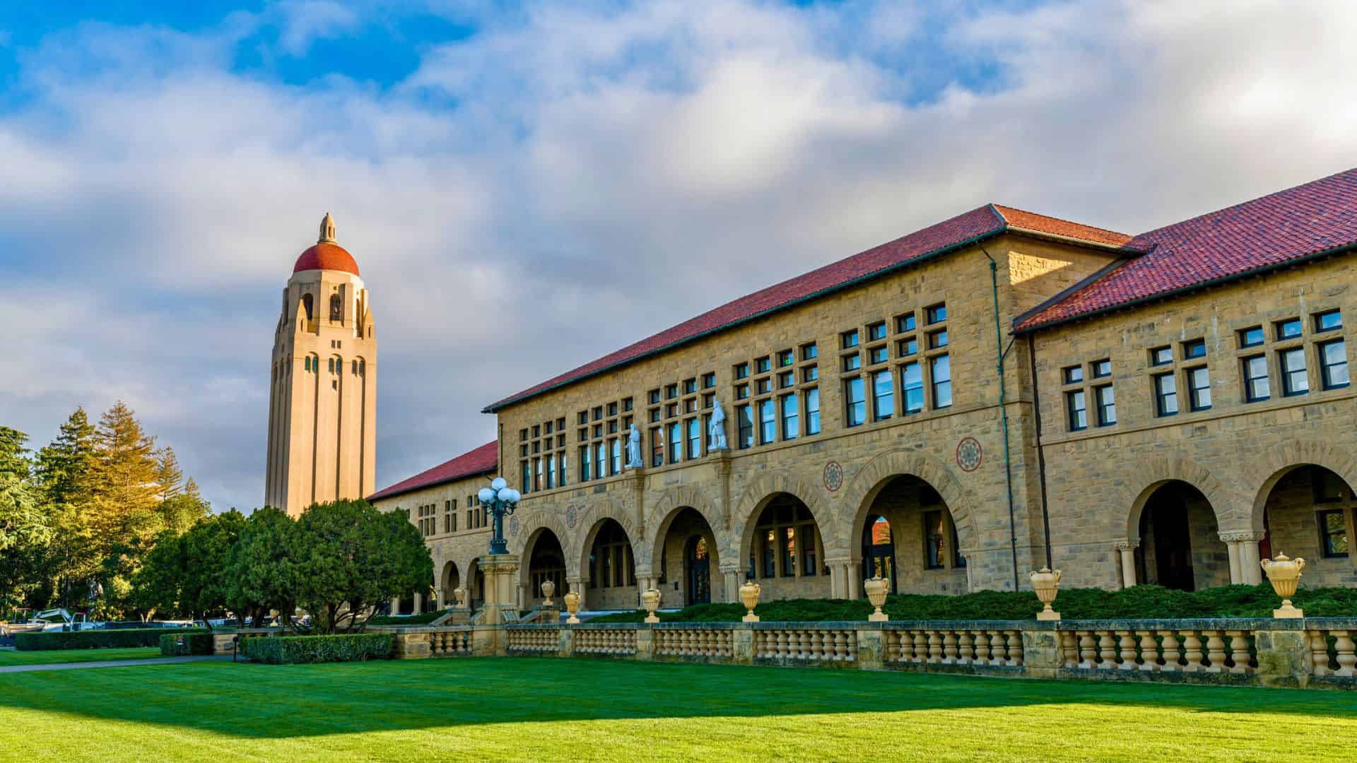 Historic stone building with arched windows and red roof, with a tall tower in the background and a green lawn.