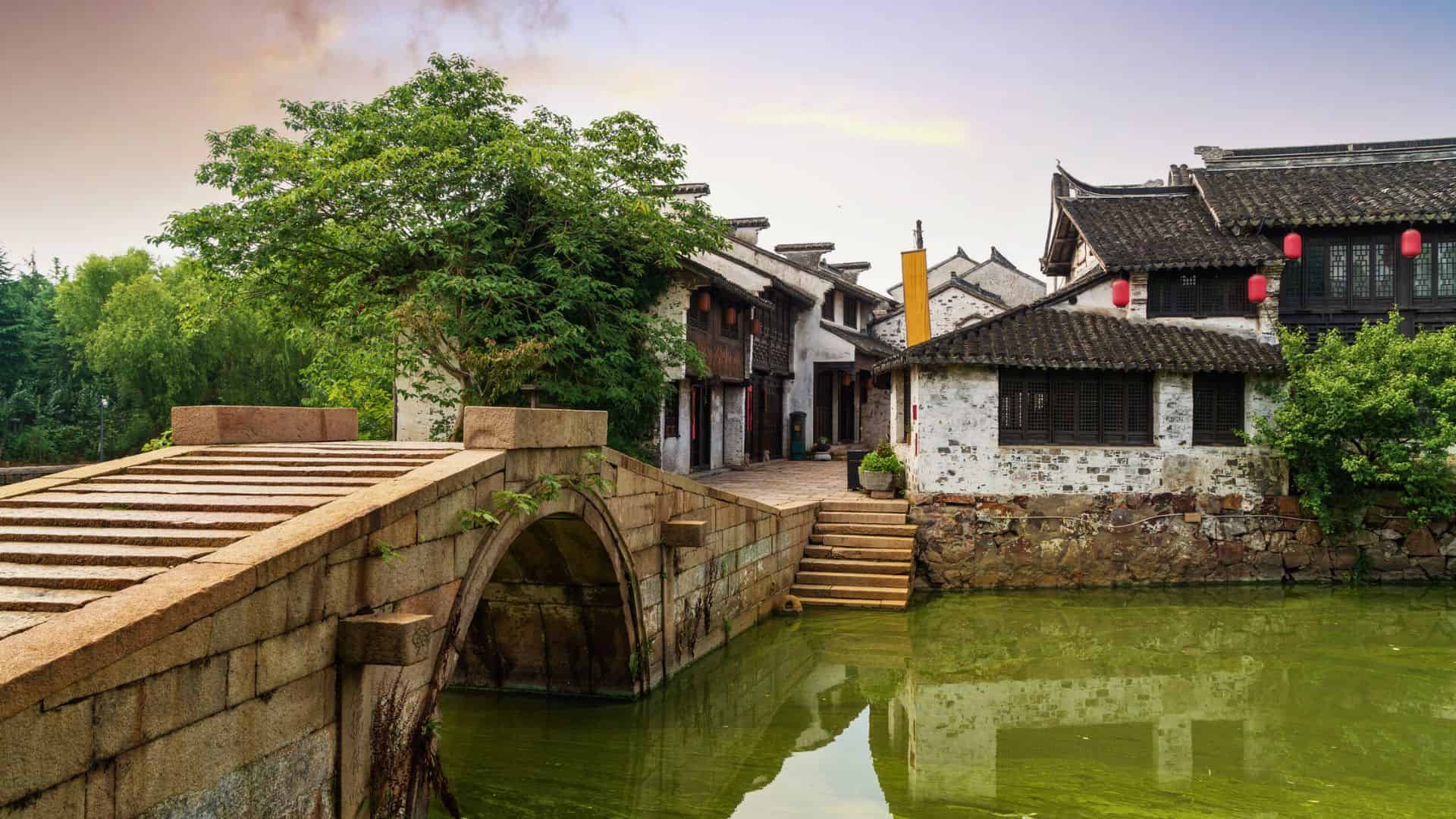 Stone bridge over a green canal, beside traditional Chinese buildings with lanterns and lush trees.