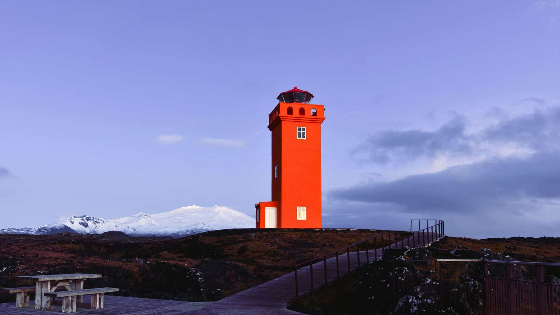 Bright orange lighthouse stands on a hill with snowy mountains and a picnic table in the background.