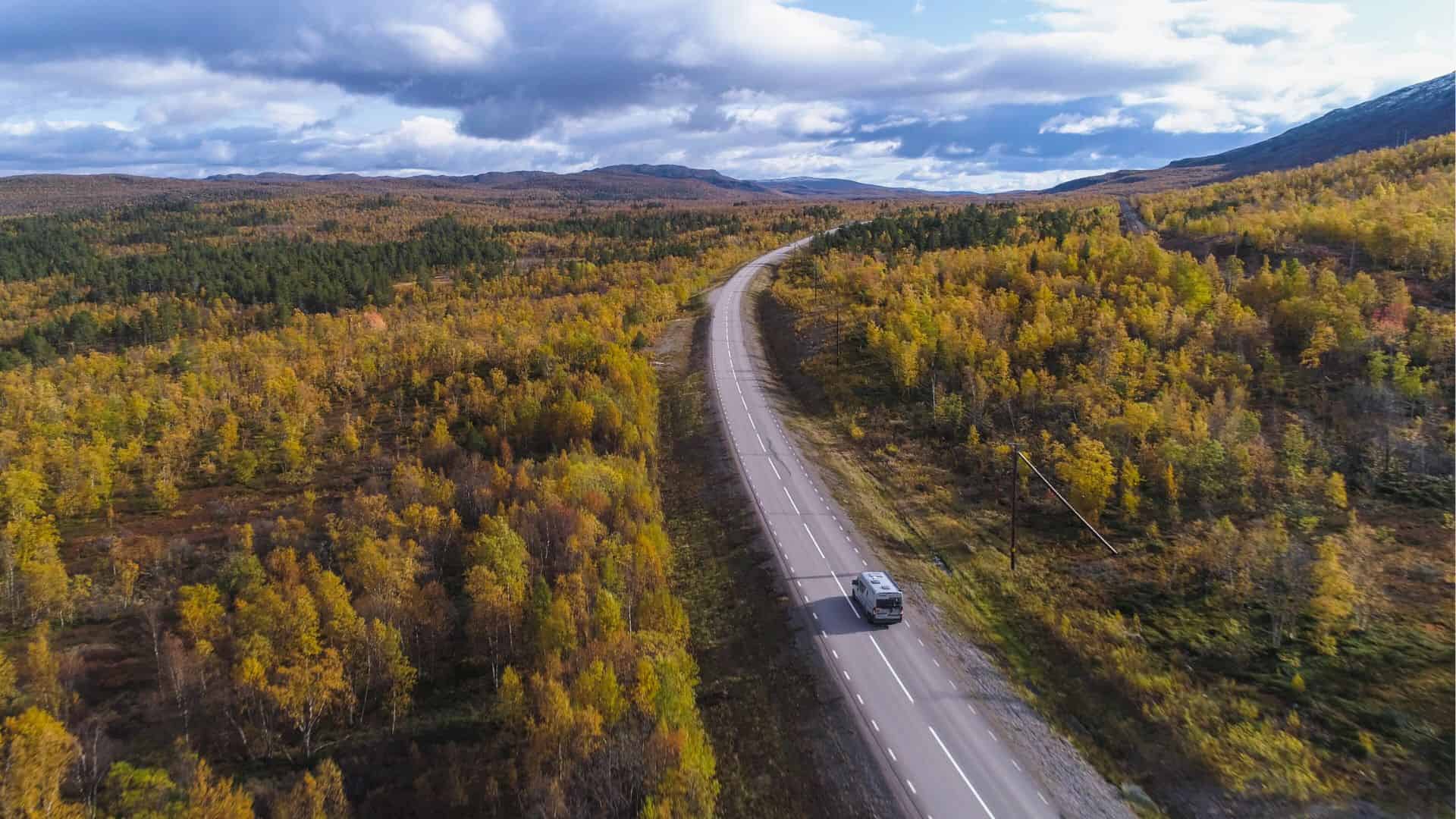 A car drives on a winding road through a forest with autumn foliage under a partly cloudy sky.