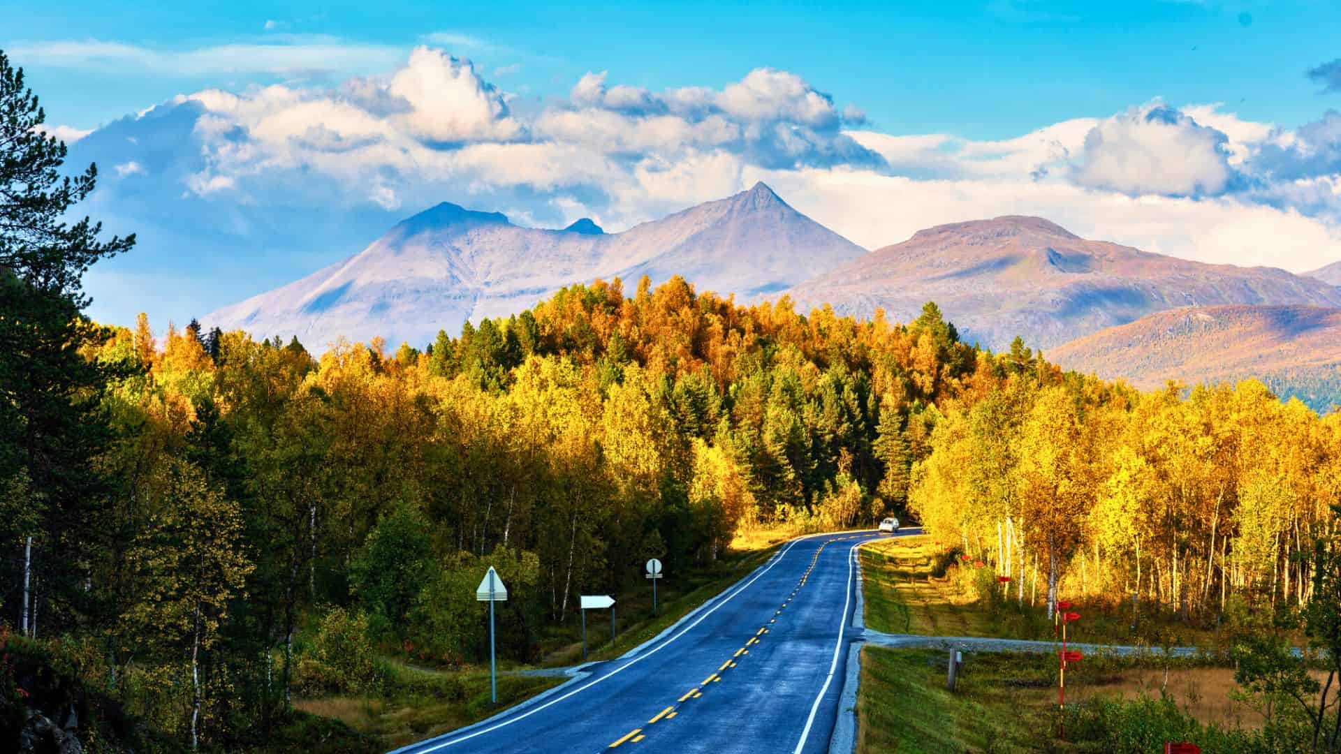 A winding road leads through autumn trees toward distant mountains under a partly cloudy blue sky.