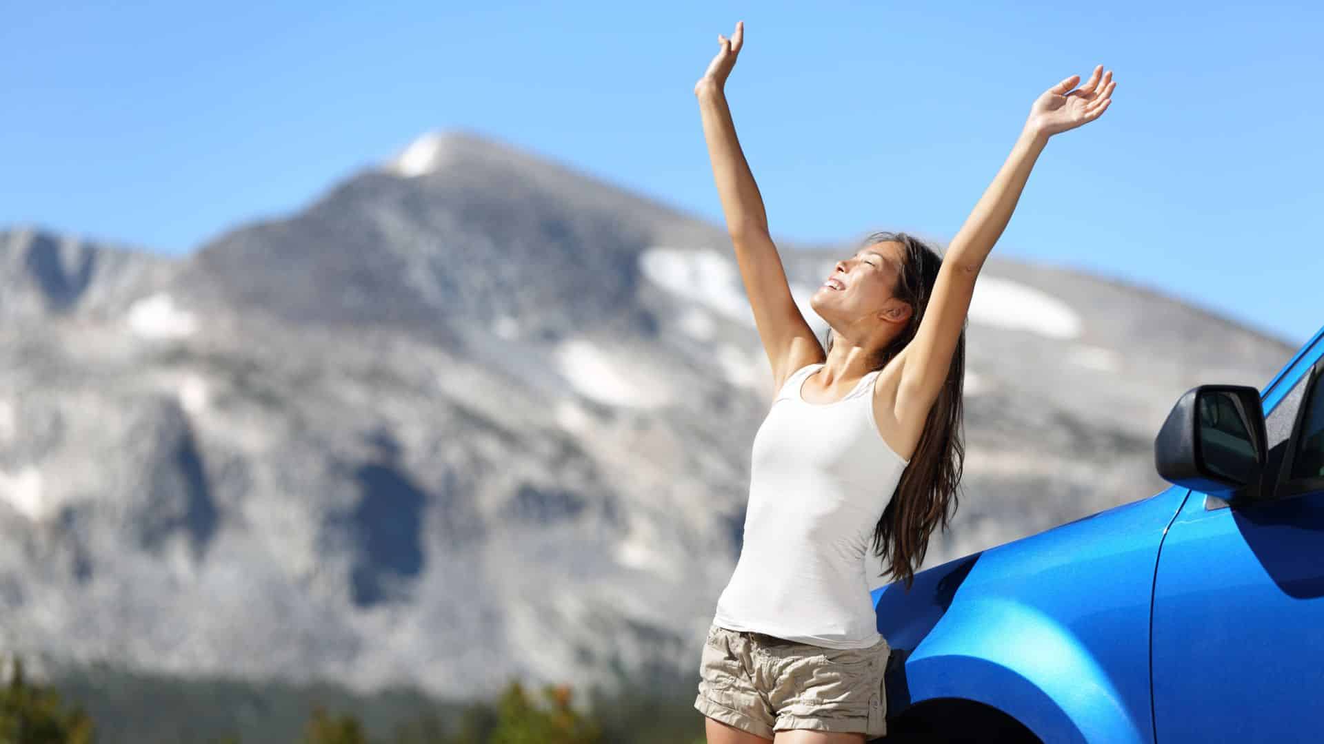 Woman smiling with arms raised, standing by a blue car with mountains in the background.