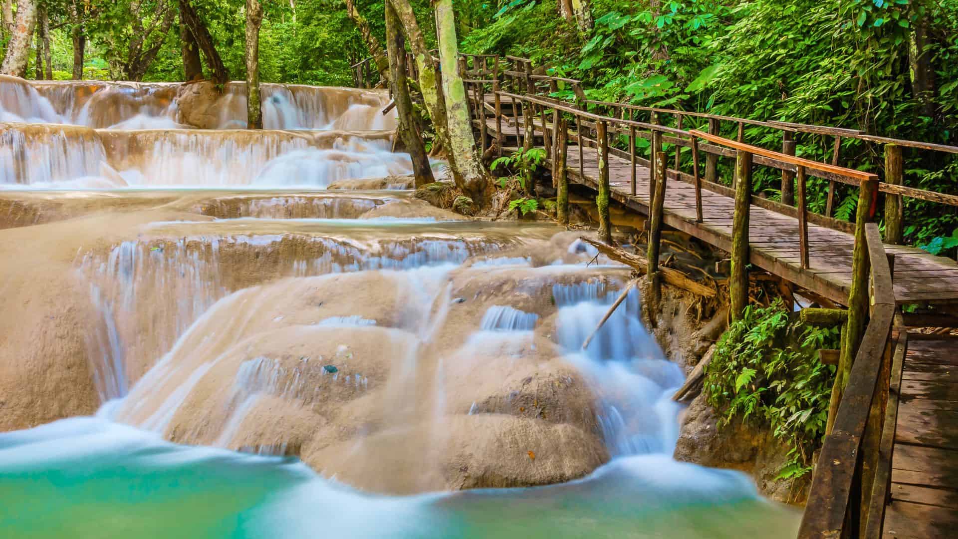 Wooden walkway beside cascading waterfalls in a lush green forest, with clear turquoise water flowing below.