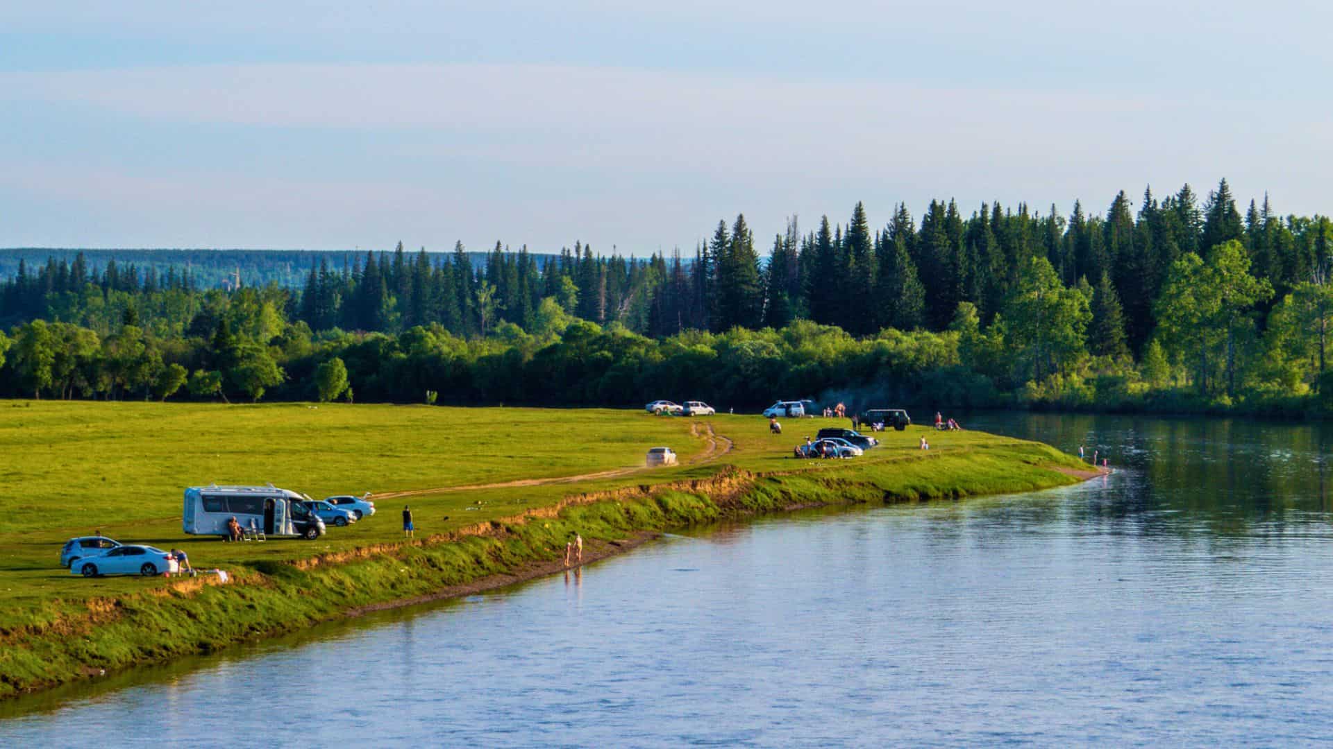 People camping and relaxing by a grassy riverbank with cars, RVs, and a forest in the background.