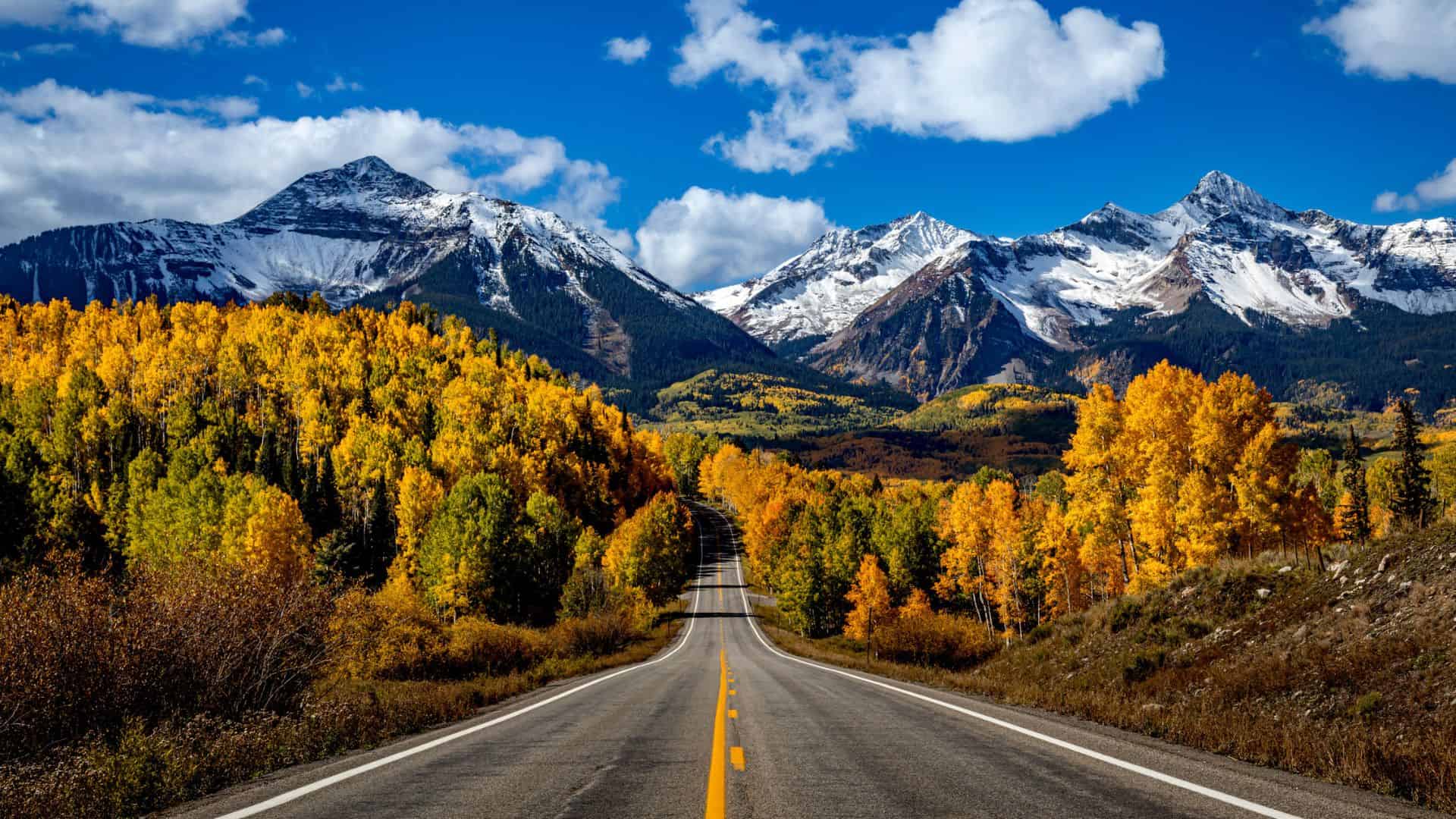 A road leads through autumn trees toward snow-capped mountains under a blue sky with scattered clouds.
