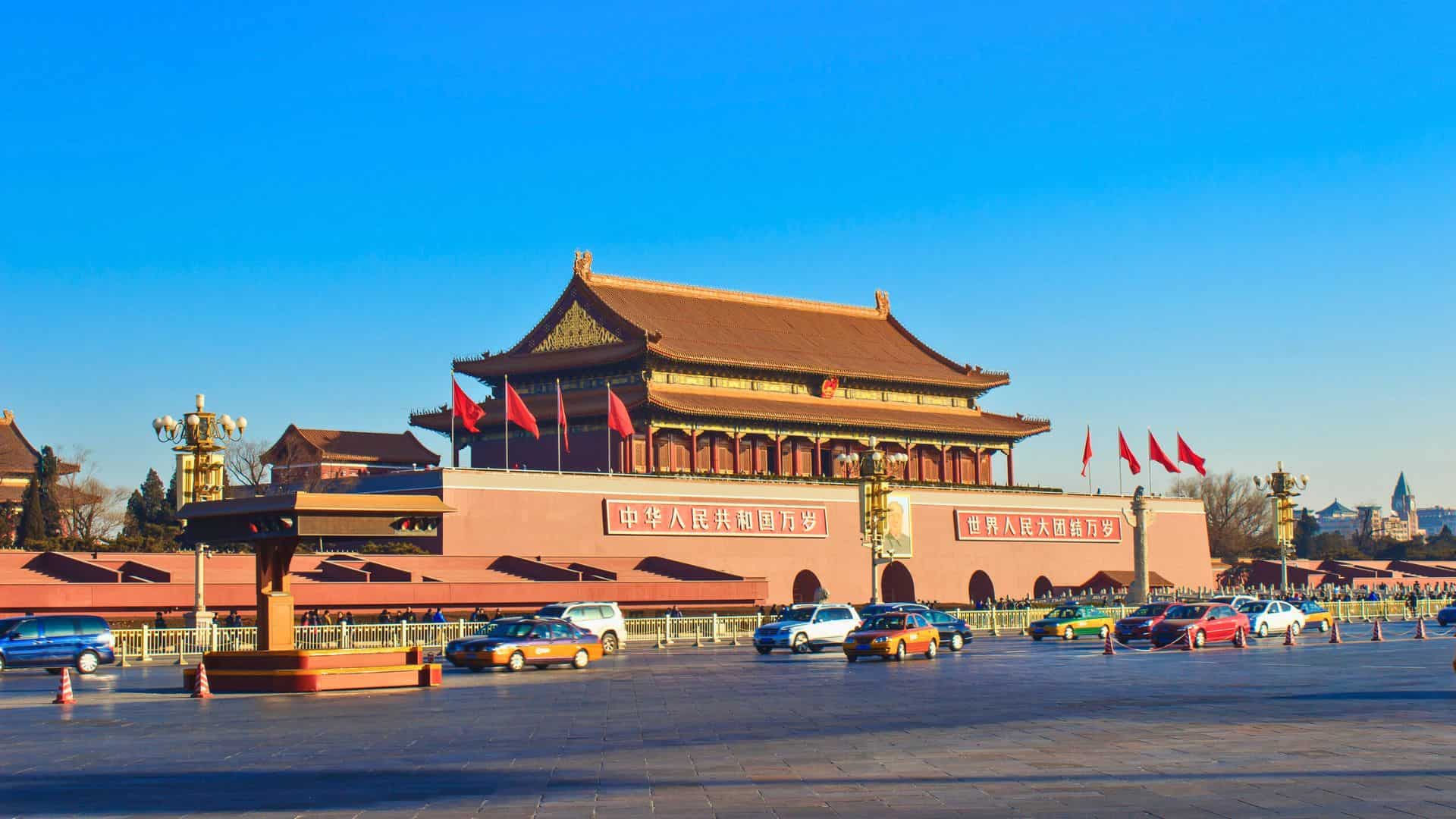 Tiananmen Gate in Beijing, China, with red flags and cars passing in front under a clear blue sky.