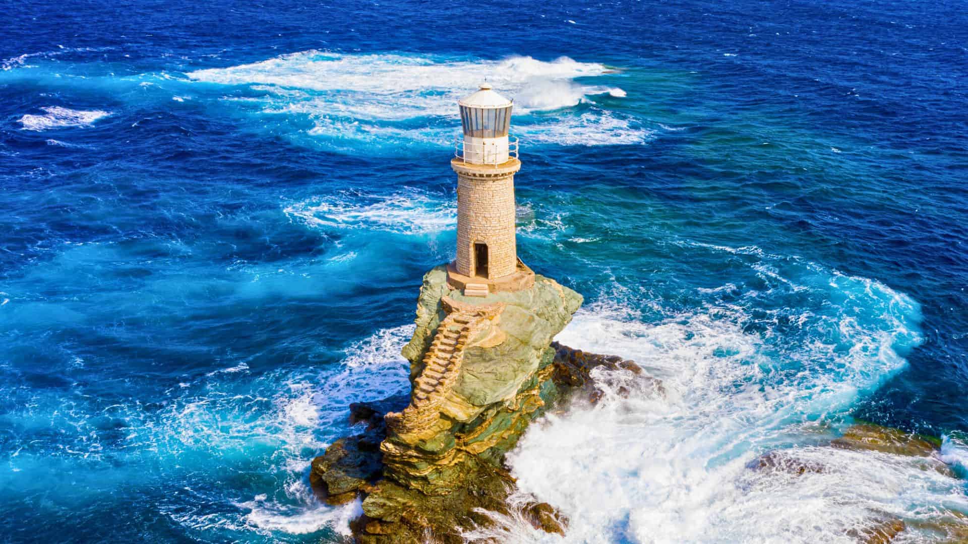 A stone lighthouse stands on a rocky outcrop surrounded by vivid blue, turbulent ocean waves.