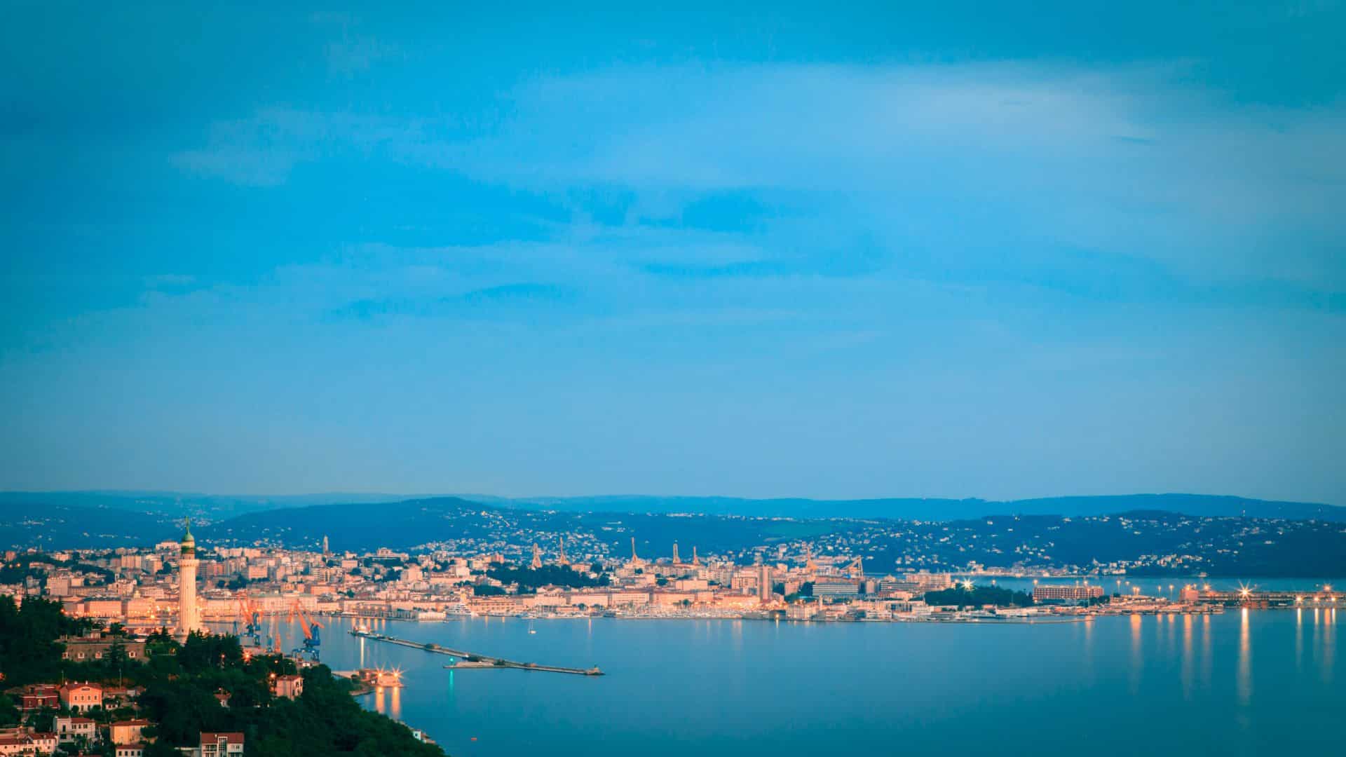 A coastal city with buildings and lights reflected on calm water under a bright blue sky at dusk.