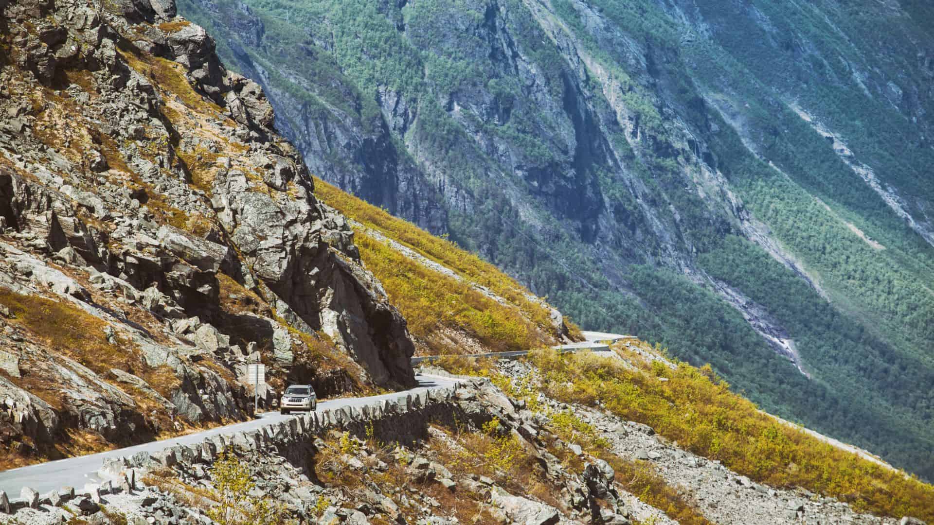 A car drives along a narrow mountain road with steep rocky cliffs and green slopes in the background.
