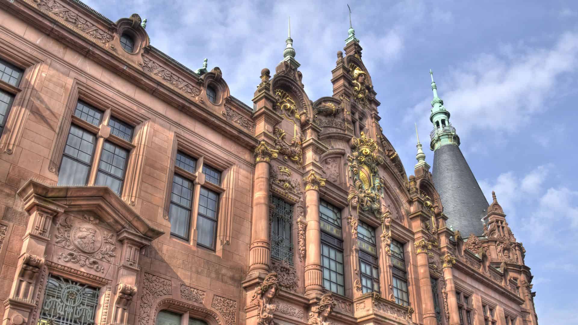 Ornate historic building with detailed stonework, large windows, and a tall spire under a blue sky.
