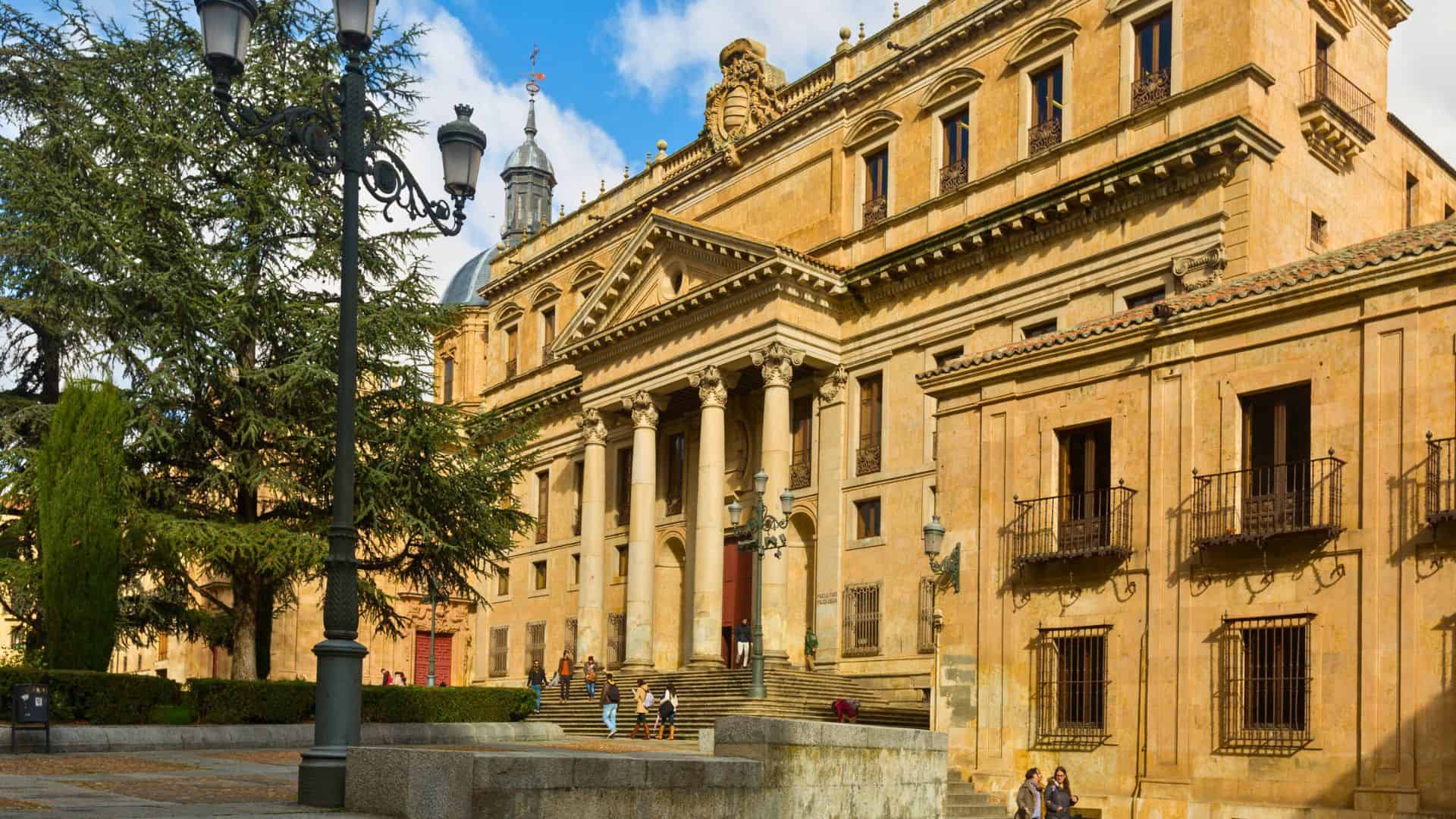 Historic sandstone building with columns and steps, people sitting outside, trees and lampposts in front.