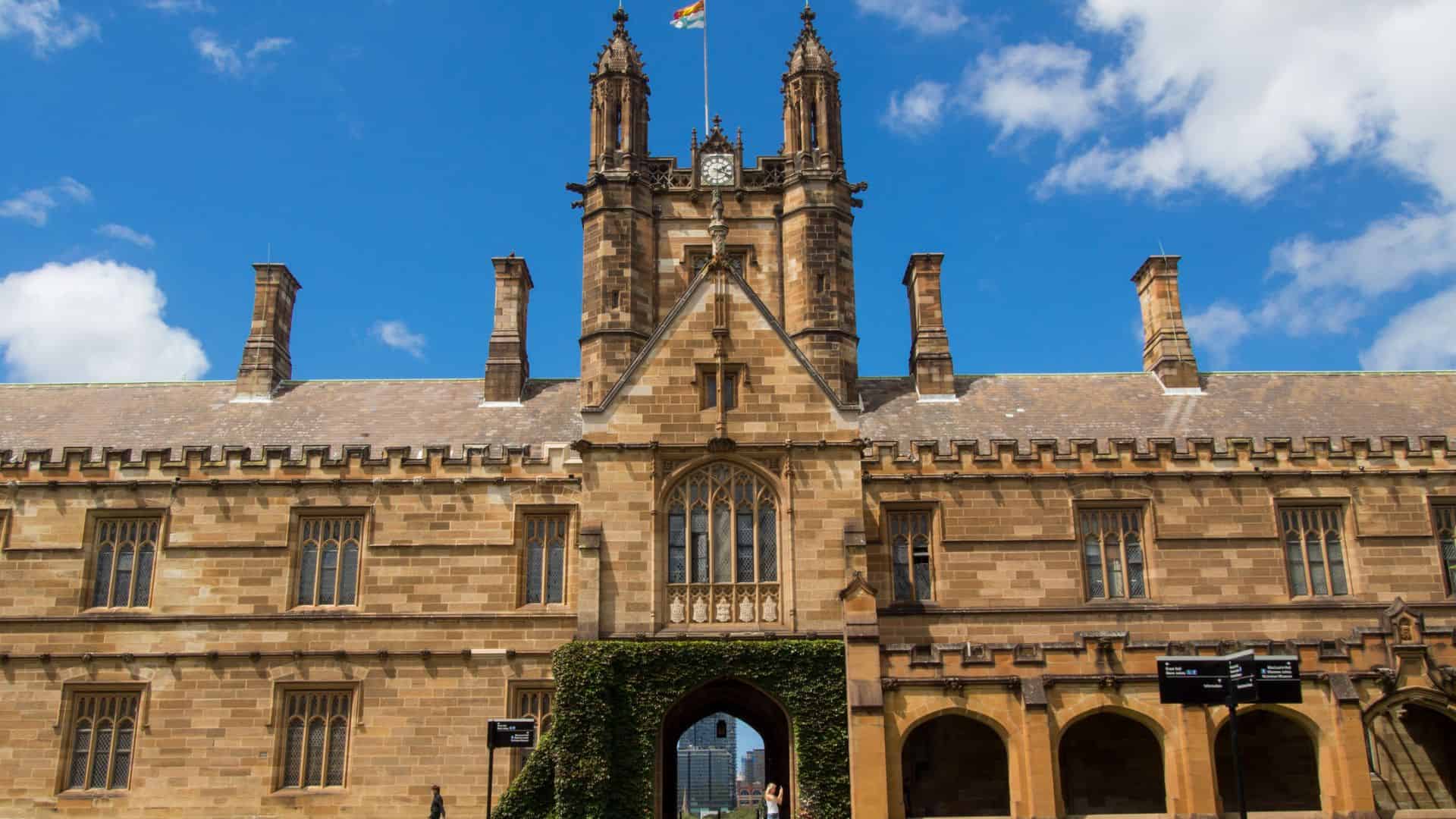 Historic sandstone university building with clock tower, archway, and blue sky with clouds above.