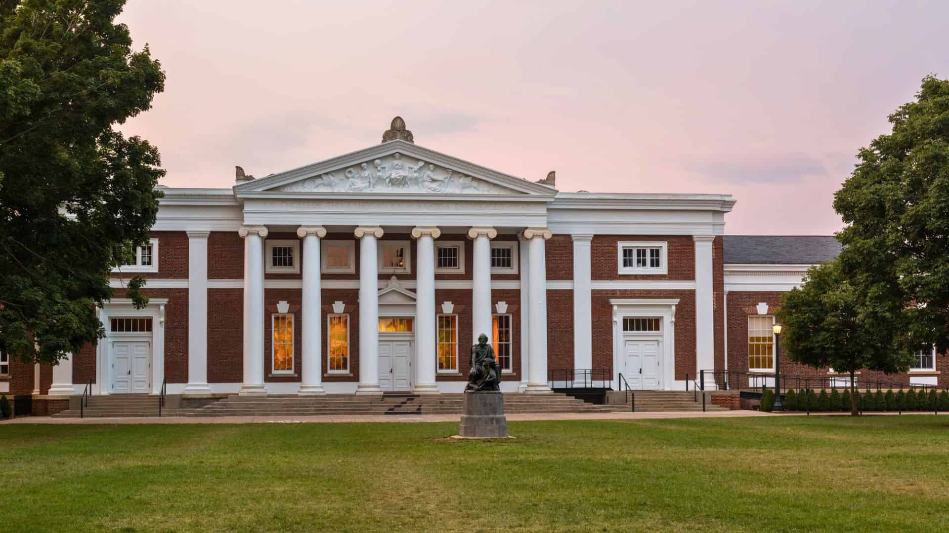 A red-brick, columned academic building with a statue on a grassy lawn at sunset.