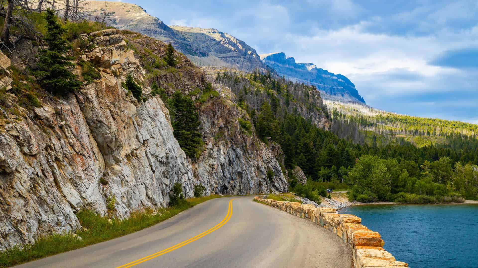 A winding road curves beside a rocky cliff, forest, and blue lake under a partly cloudy sky.