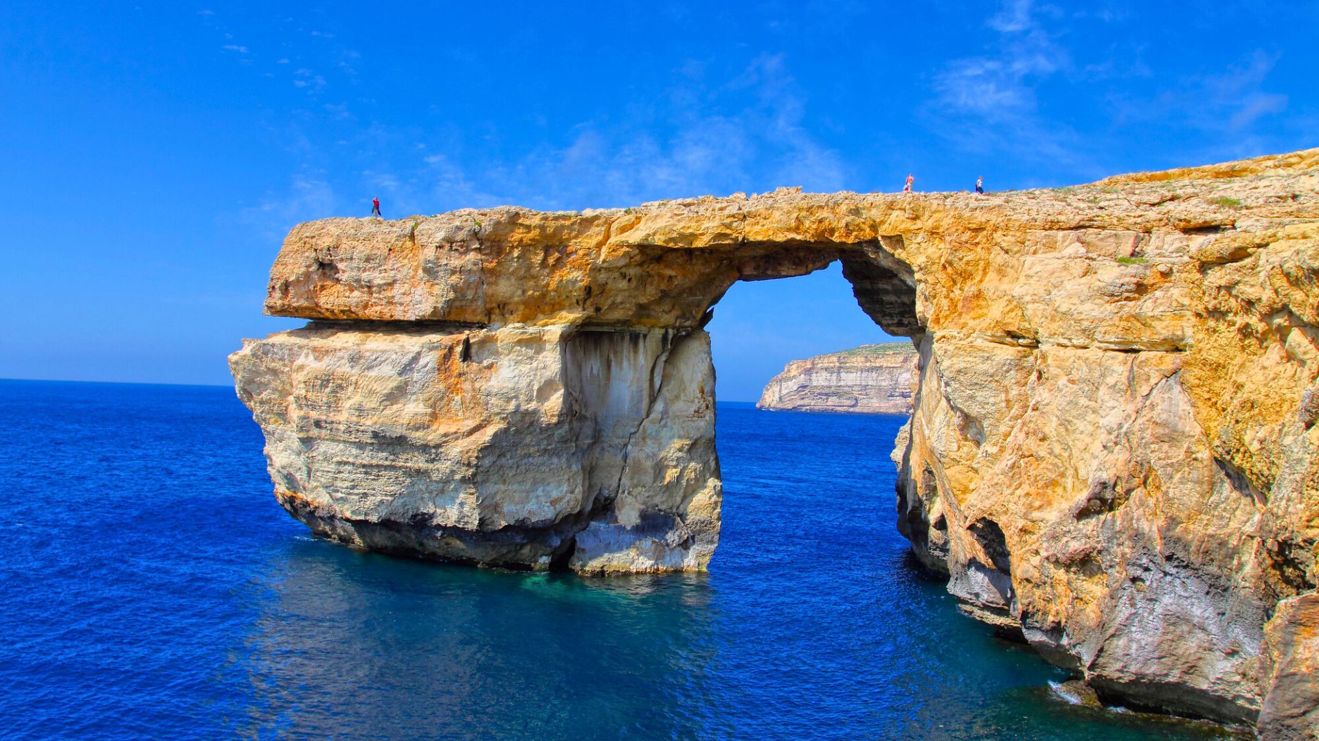 A natural rock arch rises above bright blue sea under a clear sky, with people standing on top.